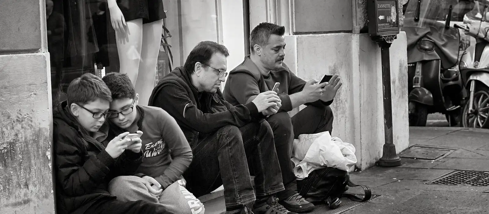 Photo noir et blanc. Deux enfants et deux hommes adultes sont assis au pied d'une vitrine, dans la rue. Tous quatre sont totalement absorbés par leur smartphone. 
